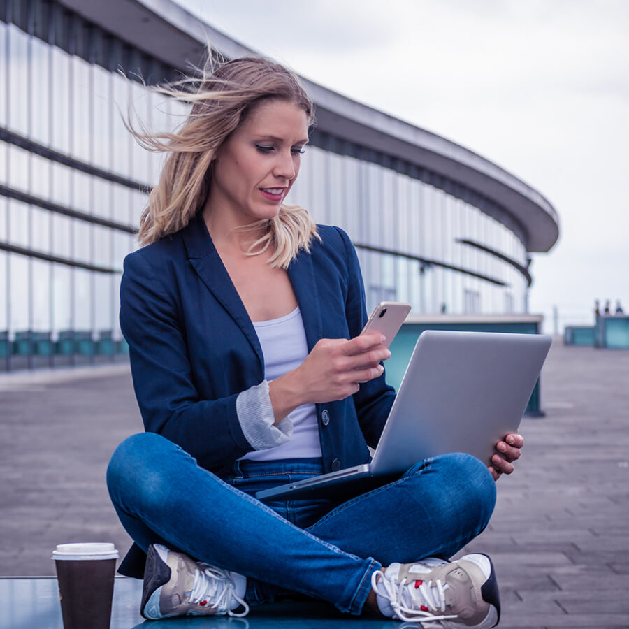 young woman, sitting outside, in front of a glass building, takes a picture with her mobile phone, on her laptop screen. The wind blows his blonde hair away.
