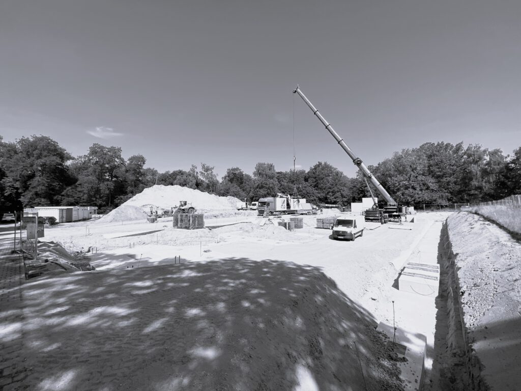 Schwarz-Weißbild von einer Baustelle, mit einem Kran und einem Auto. Die Fläche ist noch ohne Beton, mit Schotter aufgefüllt.